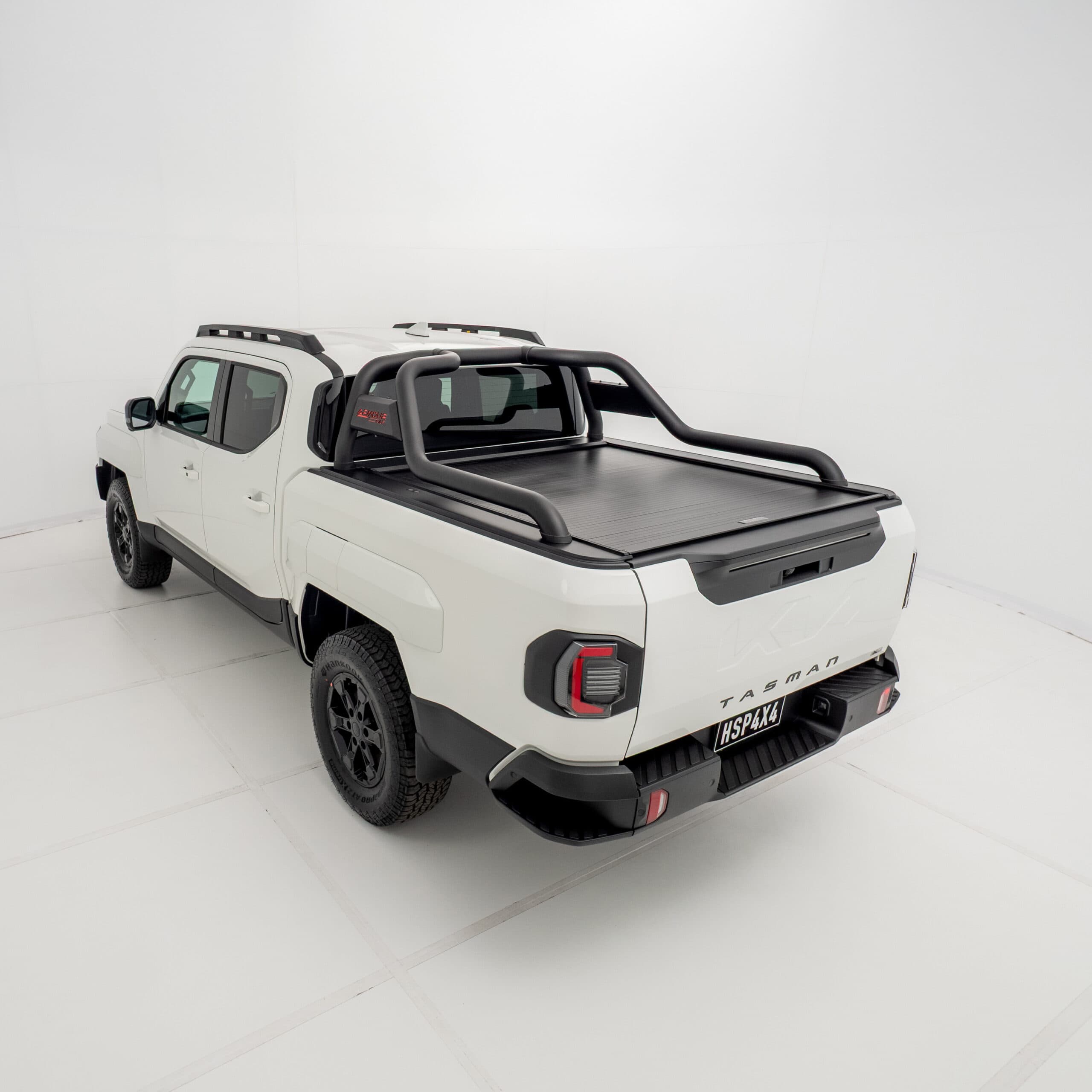 Angled 3/4 rear view of a white Kia Tasman fitted with an HSP Roll R electric roller cover in the closed position with black sports bars mounted.