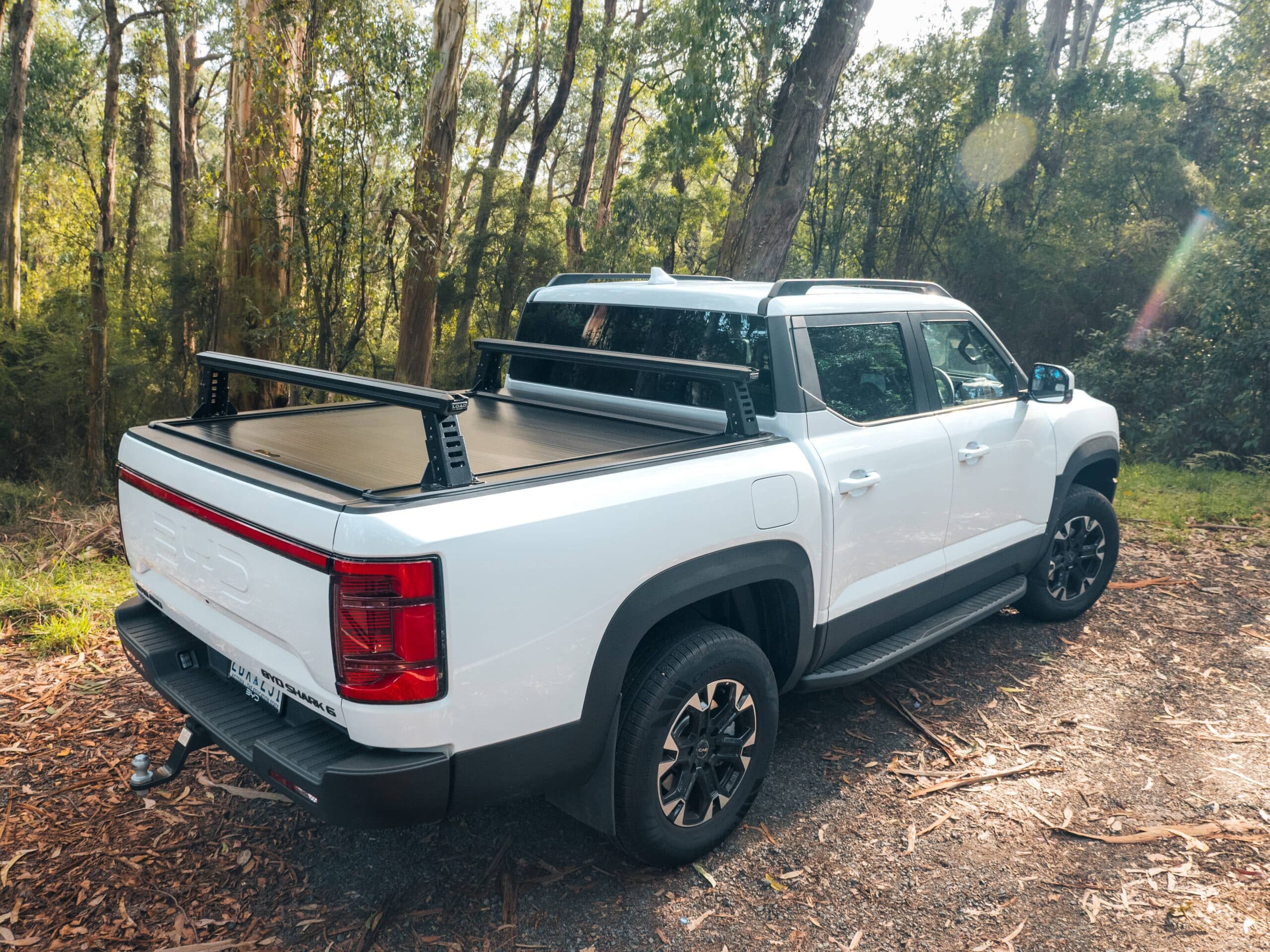Rear-side view of a white BYD Shark 6 ute parked in bushland, fitted with HSP’s Load Rack JNR system. The ute has two low-profile black rack bars mounted over a closed Roll R cover, with the tub and surrounding trees clearly visible.
